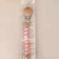 A BPA-free Pacifier Clip - Pink and Blue with wooden and food-grade silicone beads, attached to a card with product details, displayed against a plain beige background.