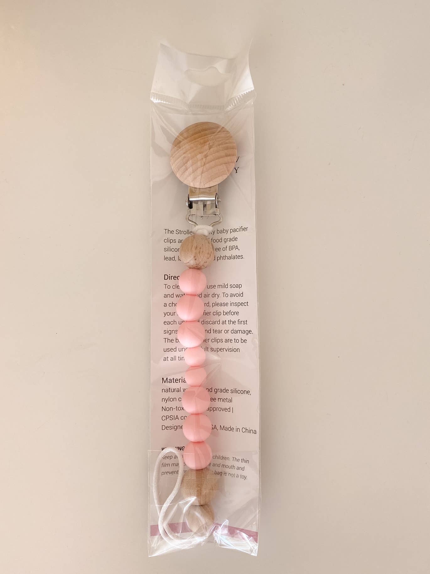 A BPA-free Pacifier Clip - Pink and Blue with wooden and food-grade silicone beads, attached to a card with product details, displayed against a plain beige background.