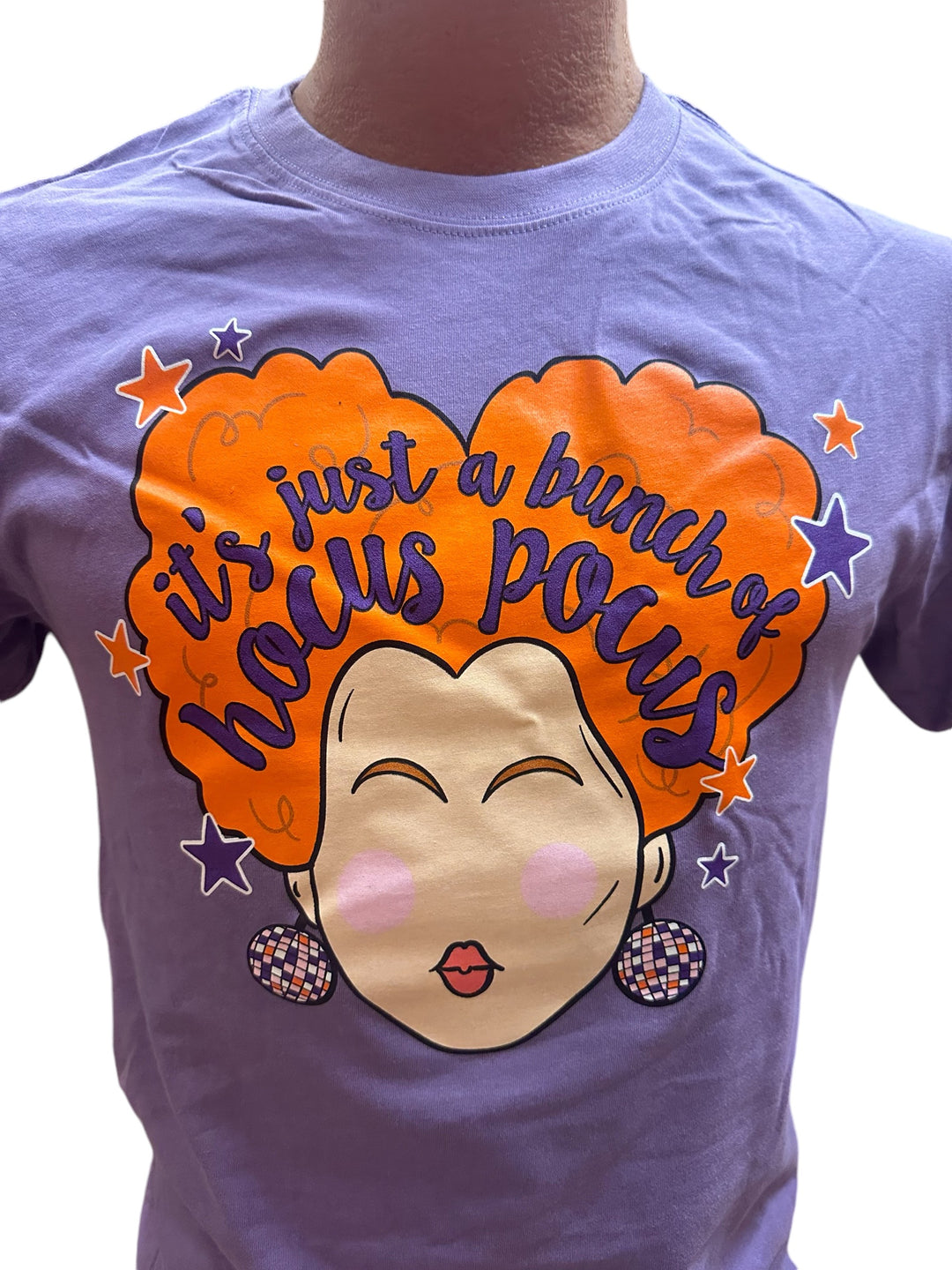 Purple “It’s Just a Bunch of Hocus Pocus” T-shirt with an illustrated woman with big orange hair, earrings, and the phrase surrounded by stars. Available in sizes S-XXL.