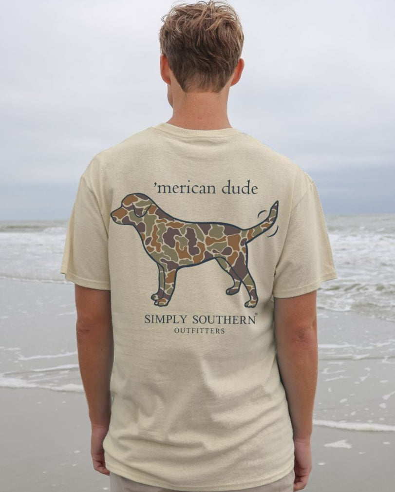 Rear view of a man wearing a beige short sleeve shirt featuring a graphic of Camo Dog outlined map and text "'merican dude - Simply Southern," standing on a sandy beach with overcast skies.