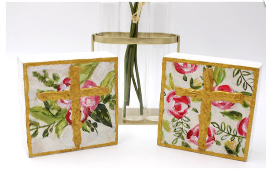 The product is a set of two hand-painted wood block signs, each measuring 4x4. The sign features a floral cross design and can also function as candle holders. The colors used in the painting are vibrant and clear, providing an attractive aesthetic element for any indoor space.