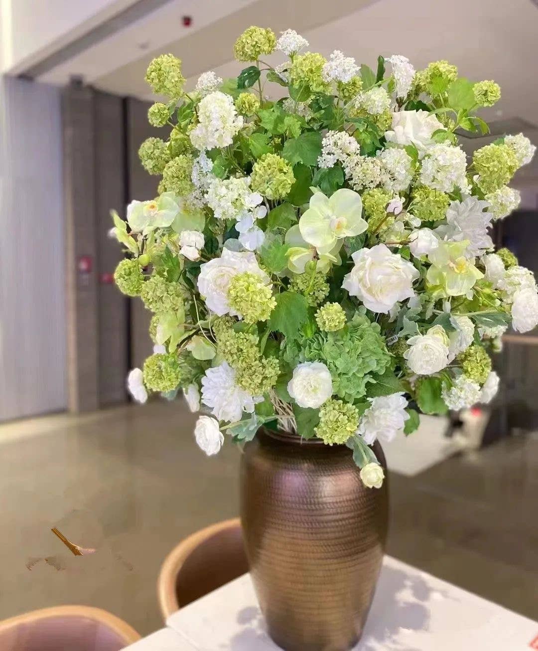 Tall, gold vase with lush green and white artificial blooms: includes Premium Faux Snowball hydrangea spray (40" long, 5 blooms), green foliage, snowball flowers, and roses. Placed on a table.