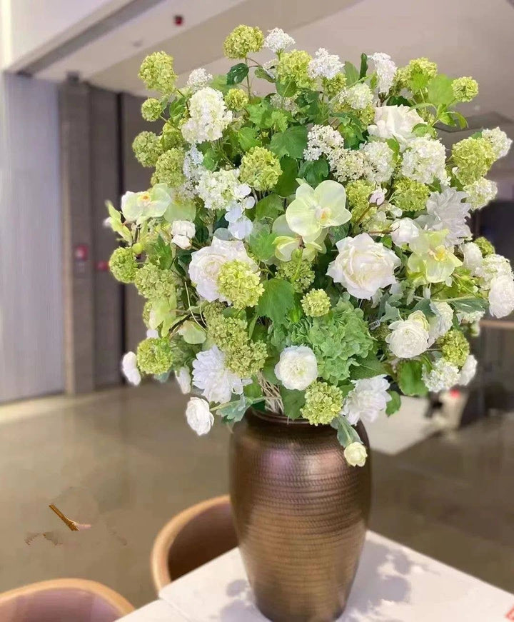 Tall, gold vase with lush green and white artificial blooms: includes Premium Faux Snowball hydrangea spray (40" long, 5 blooms), green foliage, snowball flowers, and roses. Placed on a table.