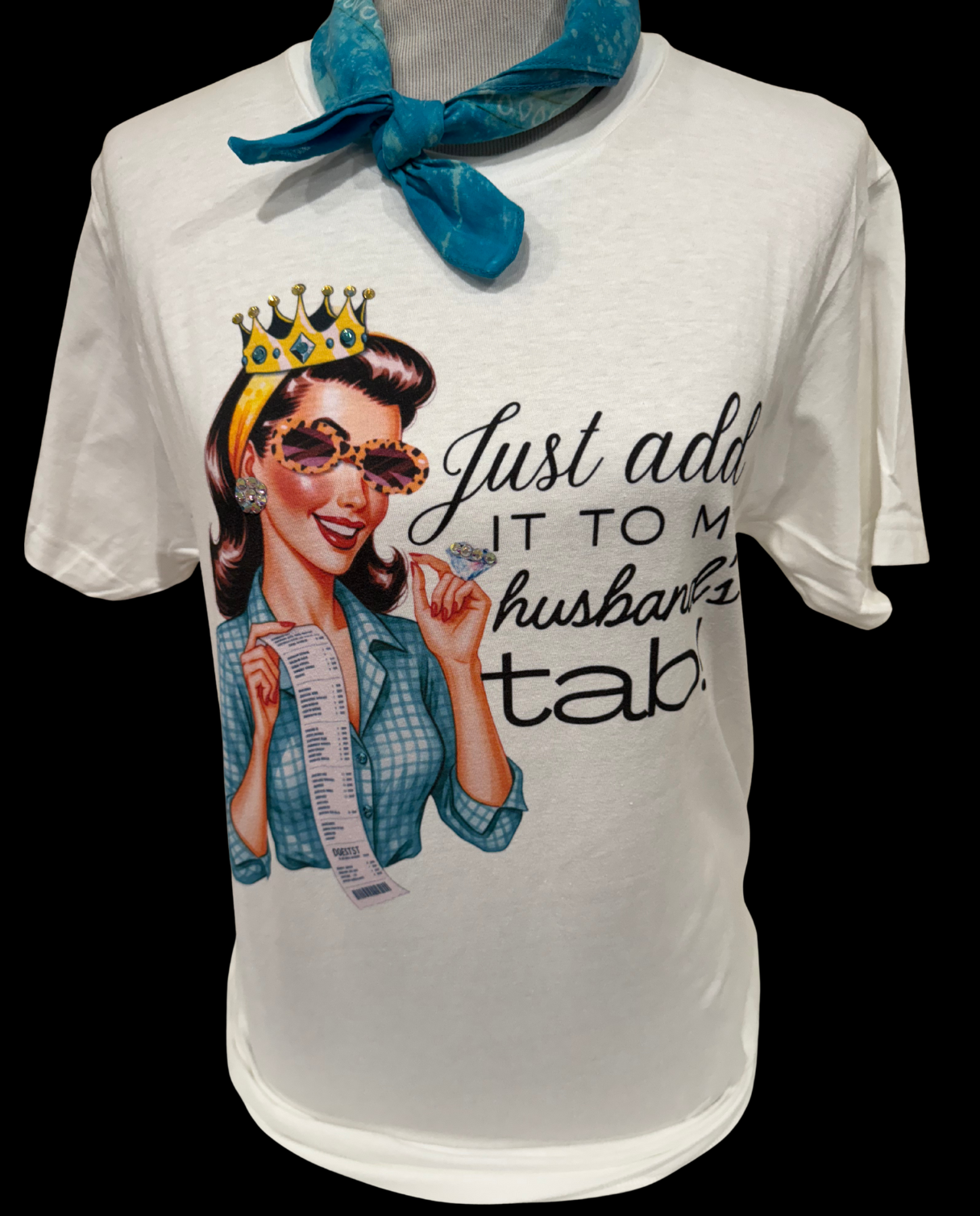 White crew-neck T-shirt with a retro-style woman in a crown and sunglasses holding an extra-long receipt, featuring the text: “Just add it to my husband’s tab.” Available in size M.