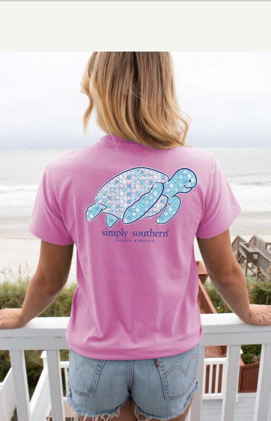 Sentence with Product Name: A person stands on a balcony facing the ocean, wearing a Preppy Turtle t-shirt featuring a blue patterned turtle design. Their blond hair is visible from the back.