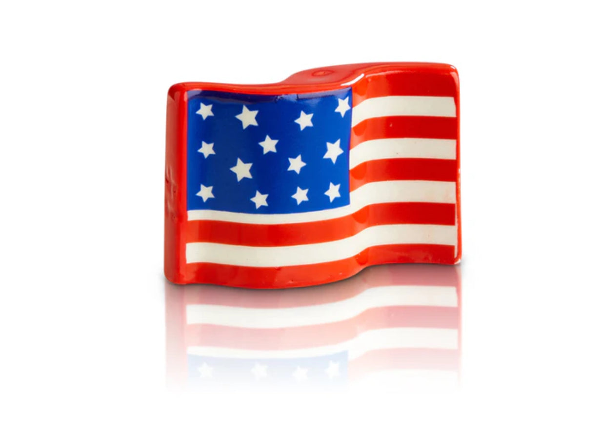 A small, wave-shaped figurine resembling the United States flag with red and white stripes and white stars on a blue background. This top-selling Nora Fleming Mini has a reflective surface that casts a mirrored reflection below it. Sizes available: Small.