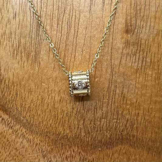 The Ezra Gold Pendant Necklace has a delicate gold chain and a textured, barrel-shaped centerpiece with a central diamond and crystal accents. Available in 16", 18", and 20" lengths.