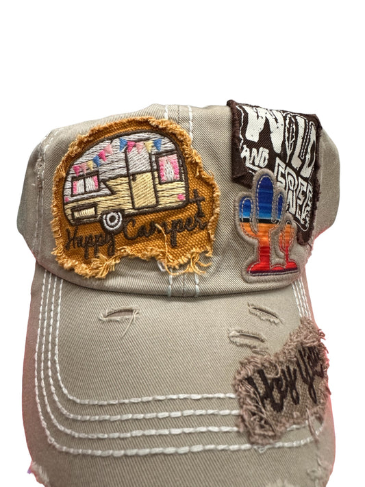 Light gray, worn-out hat with patches: a camper with "Happy Camper" text, a colorful cactus with "Wild and Free" text, and a frayed patch with "Hey y'all!" text. Visible distress marks and exposed stitching. Perfect for outdoor activities. Sizes: One size fits all.