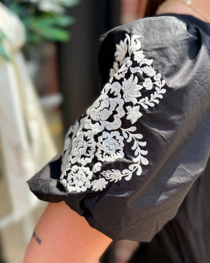 Close-up of the Tracey Aven Vines Black Dress with white floral embroidery on the sleeve and smocked waist; available in sizes XS–XL.