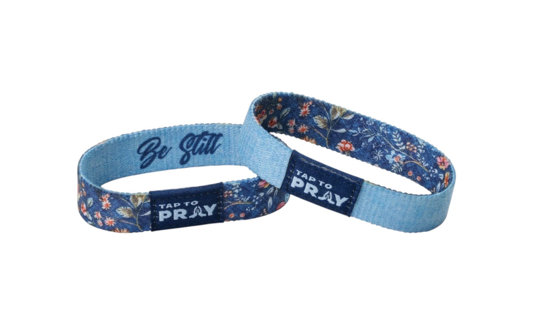Two Tap To Pray™ Wildflower wristbands with blue floral pattern, rectangular tags, and “Be Still” script inside. Available in various sizes.