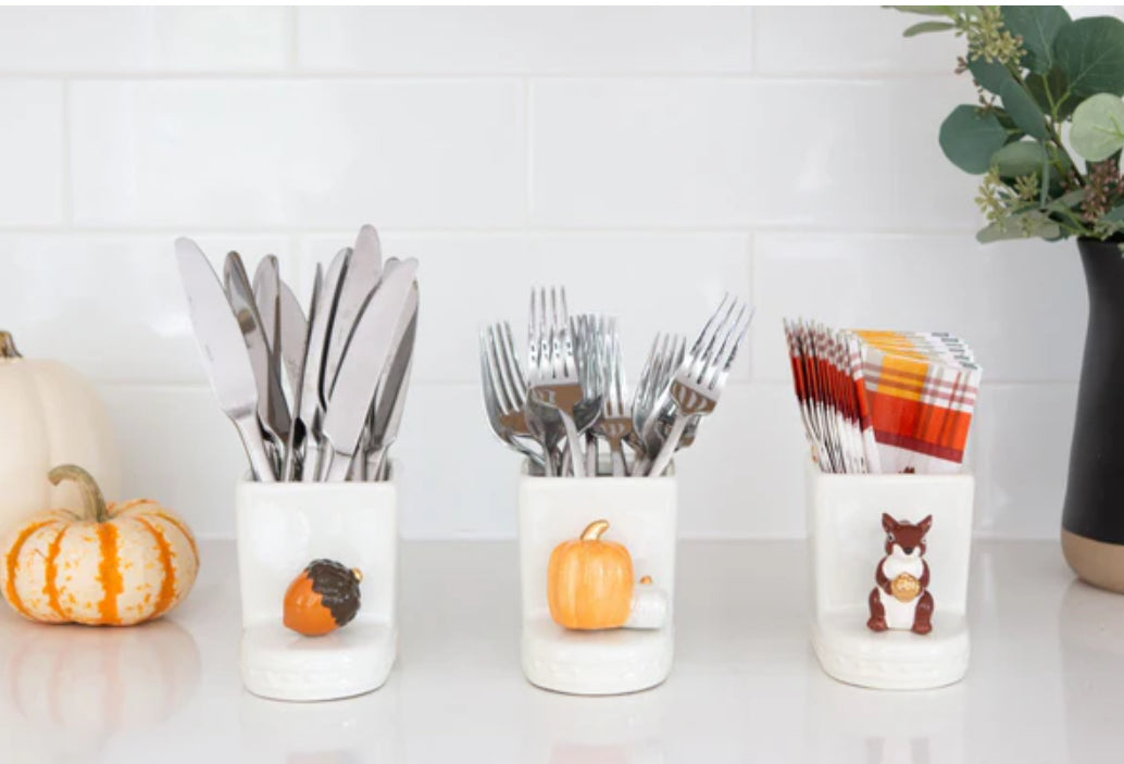 Three white ceramic containers from the Nora Fleming Minis collection, each with autumn-themed decorations. First container (acorn design) holds knives; second container (pumpkin spice motif) holds forks; third container (squirrel motif) holds napkins. Sizes available: not specified.
