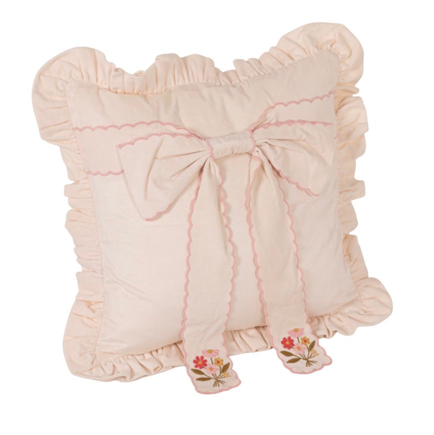 Square light cream pillow with delicate ruffled edges, featuring a large fabric bow with pink scalloped trim and embroidered pink and yellow flowers on the bow’s tails. Sizes available: 18″x18″, 20″x20″.
