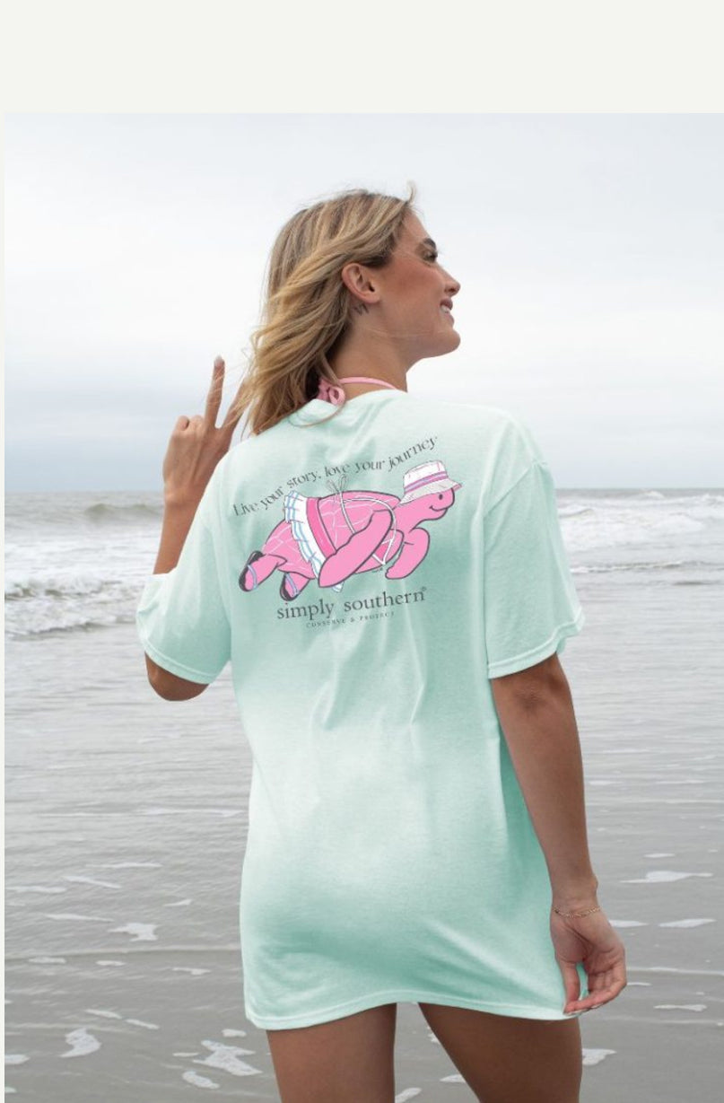 A woman on a beach wearing a BKNI-CHINCHILLA T-Shirt with a pink flamingo design and text, facing away and pointing upwards, with ocean waves in the background.