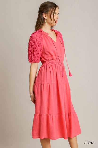 Coral Linen Midi Dress features an A-line design with textured puff sleeves, elastic waist, V-neckline, and a tasseled tie. Available sizes: XS, S, M, L, XL.