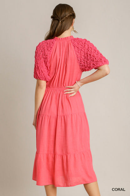 The Coral Linen Midi Dress features textured puff sleeves, an elastic waist, and a tiered A-line skirt. Available sizes: XS, S, M, L, XL.