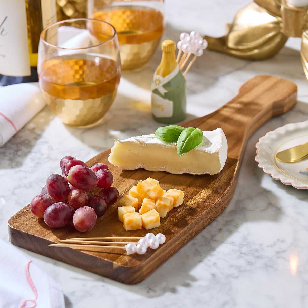 Acacia wood Champagne Toast Charcuterie Board with red grapes, cubed cheese, and a brie wedge garnished with basil. Available in small, medium, and large sizes.