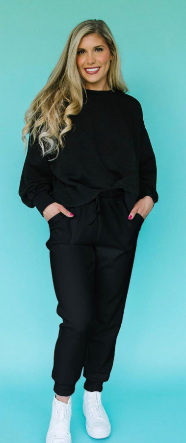 Sentence with product name: A smiling woman with long blonde hair stands against a turquoise background, wearing the Millie Lux Sweatshirt, black pants, and white sneakers. She has her hands in her pockets.