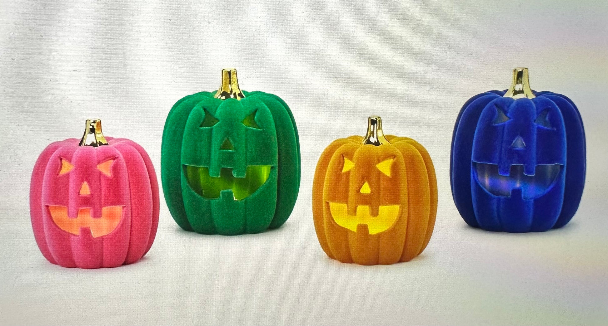 The Flocked Jack O’Lanterns come in pink, green, orange, and blue with velvet texture. Each pumpkin has carved triangular eyes, a smiling mouth, golden stems, and lights up from within. Sizes available: small, medium, large.