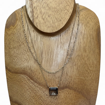 The Dula Smokey Multi-Strand Necklace features two delicate gold chains—one simple, one with a rectangular clear gemstone pendant—layered together. Available in 16" and 18" lengths.