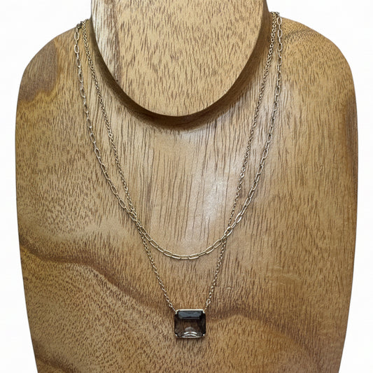 The Dula Smokey Multi-Strand Necklace features two delicate gold chains—one simple, one with a rectangular clear gemstone pendant—layered together. Available in 16" and 18" lengths.