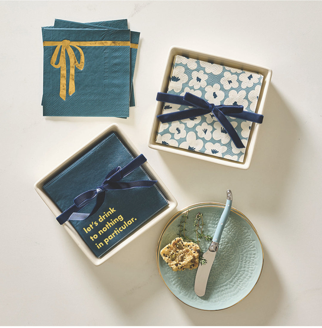 Three stacks of cocktail napkins featuring gold bow, floral, and “let’s drink to nothing in particular” prints, with velvet ribbon accents.