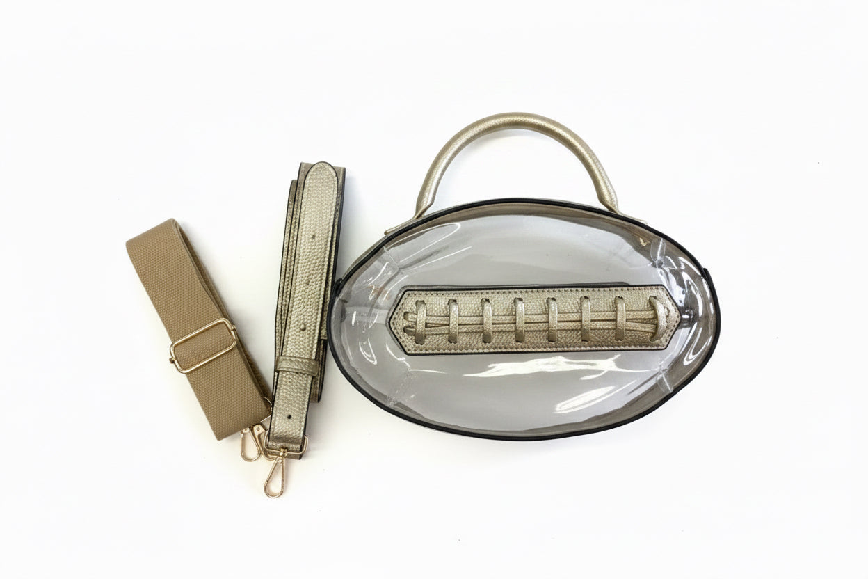 Clear oval football bag with metallic gold handles, trim, detachable strap, and interior zippered pouch. Available in one size.