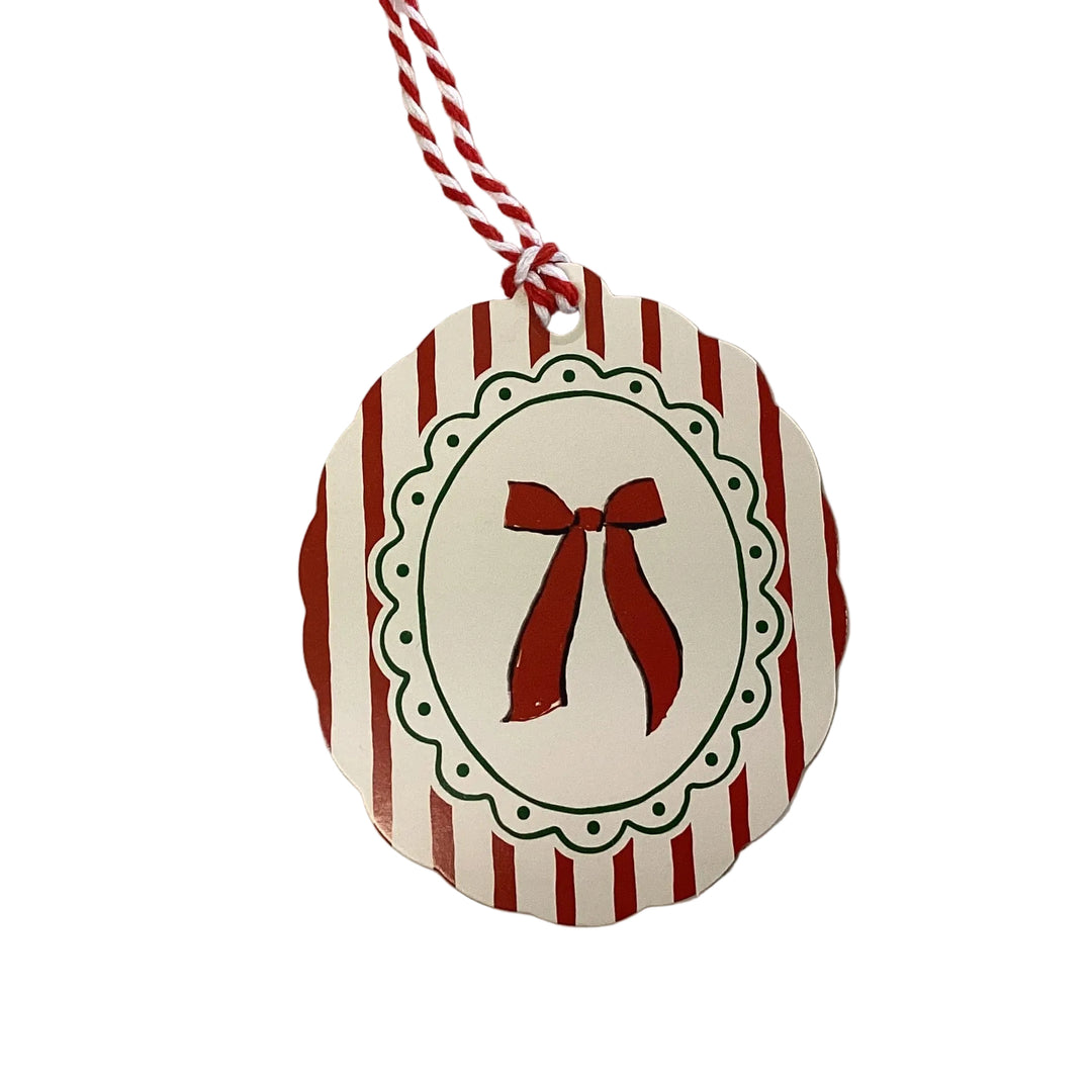 Scalloped round holiday gift tag with red and white vertical stripes, a centered red bow illustration inside a decorative green-dotted oval frame, and a red and white baker’s twine loop.