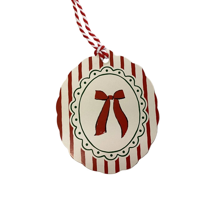 Scalloped round holiday gift tag with red and white vertical stripes, a centered red bow illustration inside a decorative green-dotted oval frame, and a red and white baker’s twine loop.