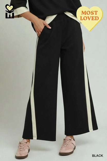 Black Colorblock Side Striped Straight Pants feature white side stripes, a wide-leg silhouette, and luxe knit fabric. Sizes available: XS-XL. Yellow heart-shaped "MOST LOVED" badge in top right corner.