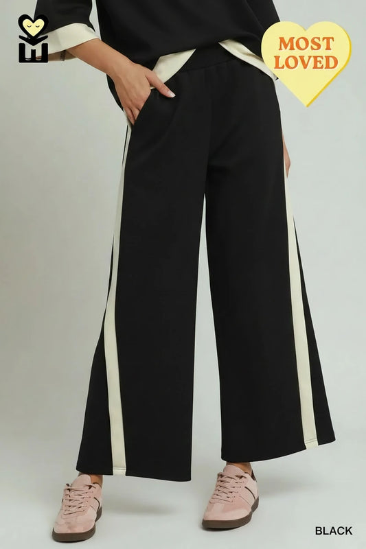 Black Colorblock Side Striped Straight Pants feature white side stripes, a wide-leg silhouette, and luxe knit fabric. Sizes available: XS-XL. Yellow heart-shaped "MOST LOVED" badge in top right corner.