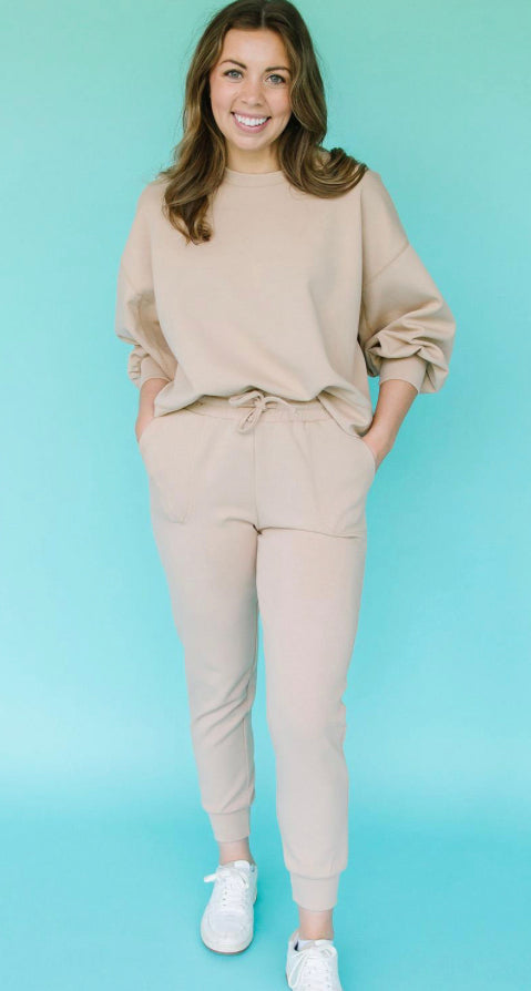 A woman in a Millie Lux Sweatshirt and coordinated sweatpants stands smiling against a teal background, hands casually in pockets. She is wearing white sneakers.