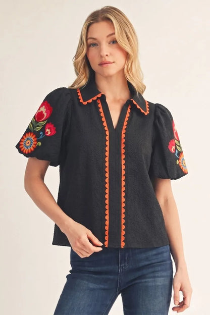 The Fall in Bloom Blouse is a black top with orange trim and vibrant floral embroidery on textured puff sleeves. Available in sizes XS-XL.