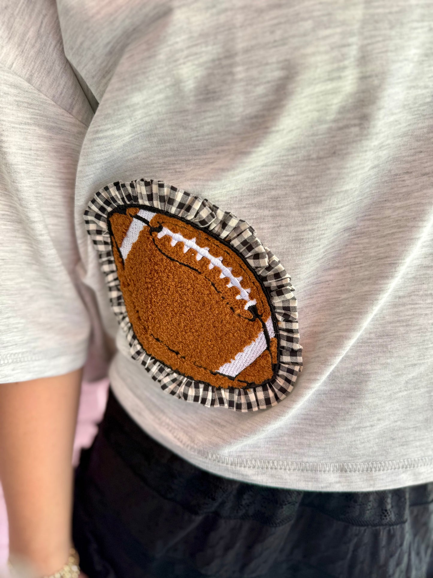 Gray oversized cropped top with a fabric football patch, white stitching, and black-and-white ruffle border near the hem. Available in sizes S-XL.