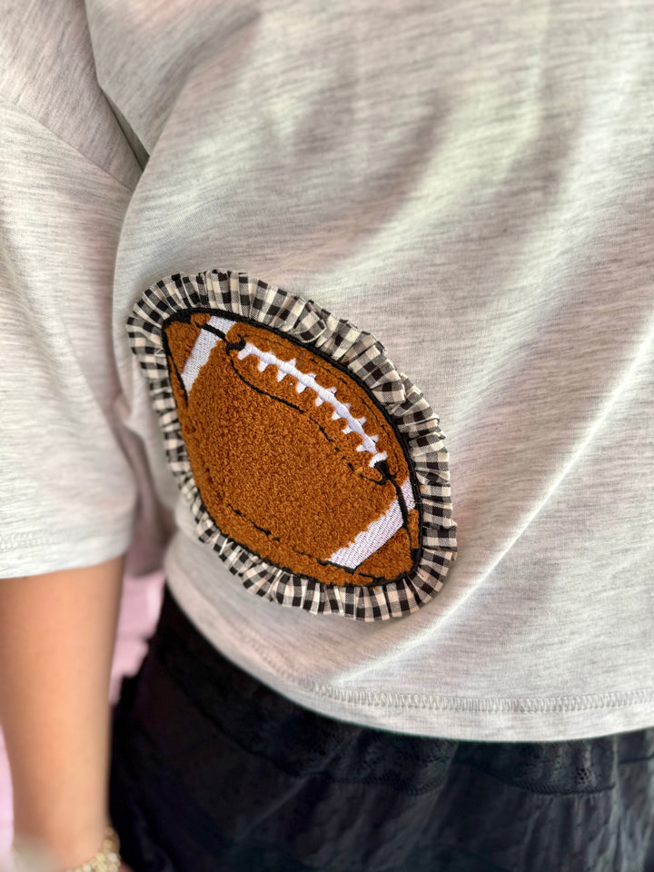 Gray oversized cropped top with a fabric football patch, white stitching, and black-and-white ruffle border near the hem. Available in sizes S-XL.