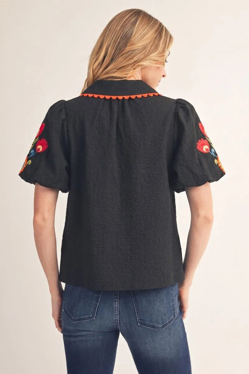 Fall in Bloom Blouse: black textured top with puff sleeves, orange scalloped collar trim, and colorful floral embroidery on sleeves. Available in sizes XS–XL.