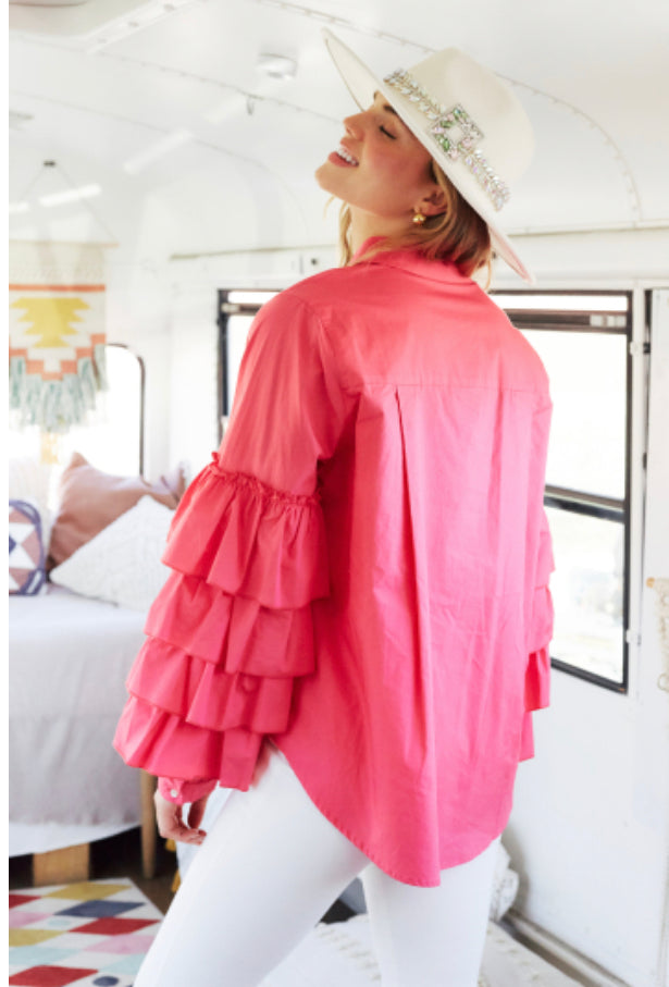 A woman in a vibrant pink Charlotte Button Down Ruffle Sleeve Top and white pants stands inside a bus turned into a cozy living space, smiling and looking away, wearing a white hat adorned with greenery.