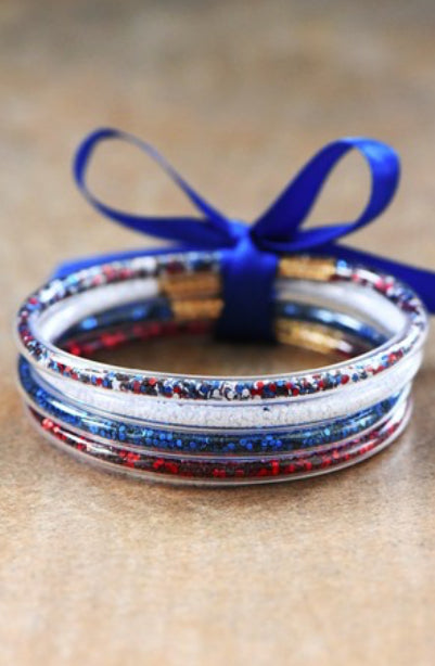 The product is a stack of Americana Patriotic Confetti Bracelet Sets. Each set comprises multiple bracelets intermingling various traditional colors including red, white, and blue. The sets come bound together by a vibrant blue ribbon for an added pop of color and convenience. They are set against a textured surface to highlight the bracelet's appeal. These bracelet sets are one size fits all.