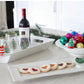 Elegant Bread Tray Stripes holding a red wine bottle and decorated wine glasses. Also includes a rectangular plate of assorted cookies and a bowl of colorful Christmas ornaments on a greenery-adorned kitchen counter. Available sizes: Standard.