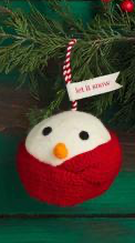 Round white felted wool Snowball Ornament with black eyes, orange nose, and red scarf, hanging by a red and white string with “let it snow” tag.