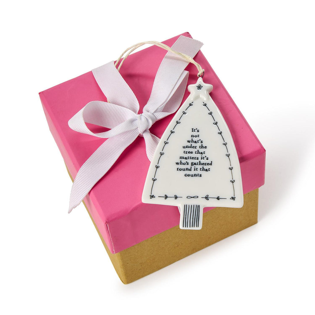 White ceramic Christmas tree hanging ornament with ribbon loop, reading, “It’s not what’s under the tree that matters it’s who’s gathered round it that counts.”.