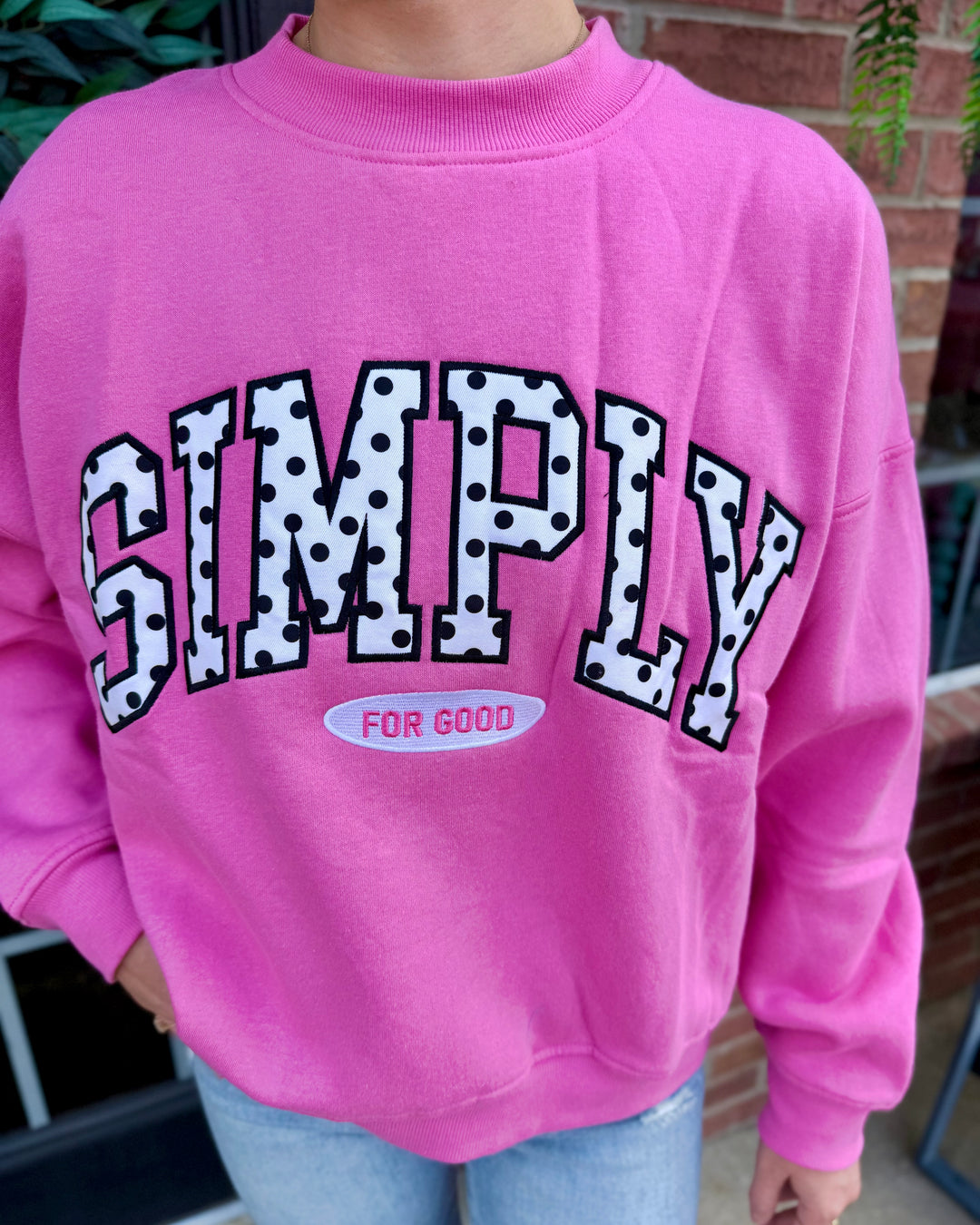 Bright pink crew neck sweatshirt with polka dots, “SIMPLY” in bold varsity lettering, and “FOR GOOD” in a small pink oval beneath. Available in sizes XS–XXL.