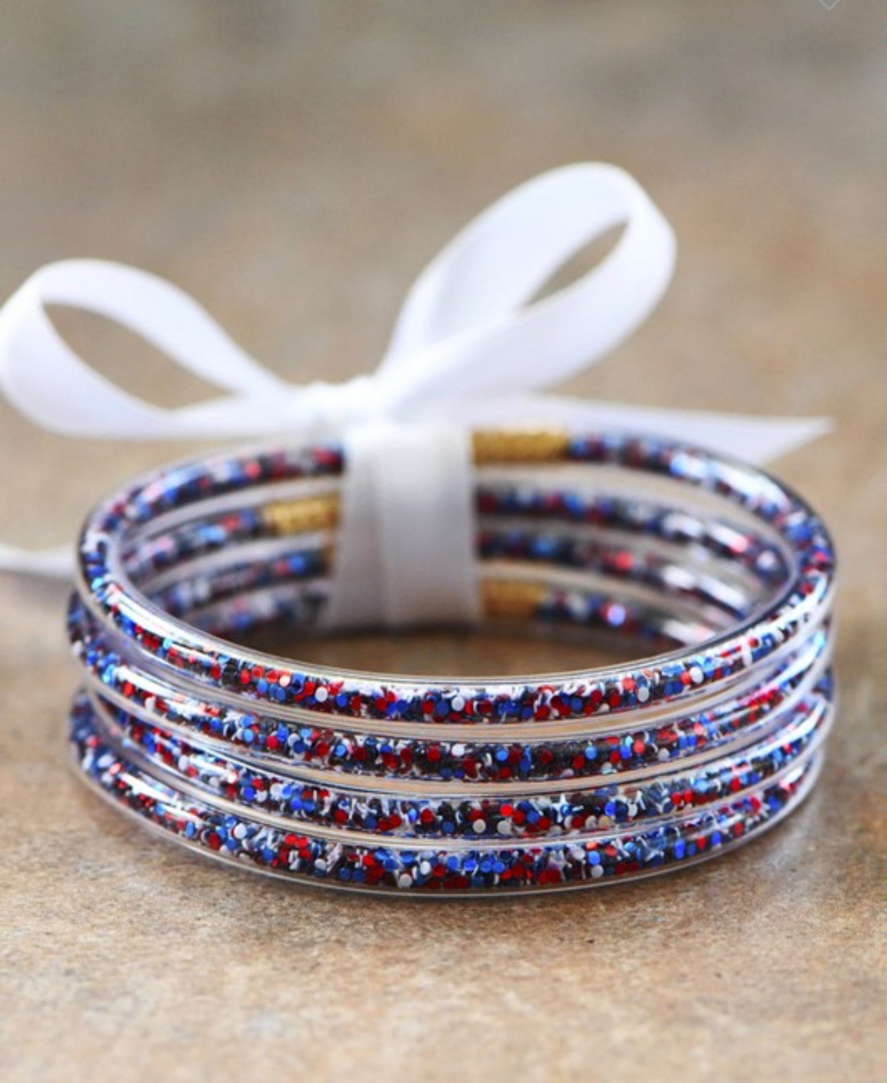 These Americana Patriotic Confetti bracelets are thin and lightweight for comfortable wear. They come in a stack, elegantly tied together with a delicate white satin ribbon. The textured surface of each bracelet reflects light beautifully, enhancing the overall look. Available in one-size-fits-all red white and blue tube bangles set of 4