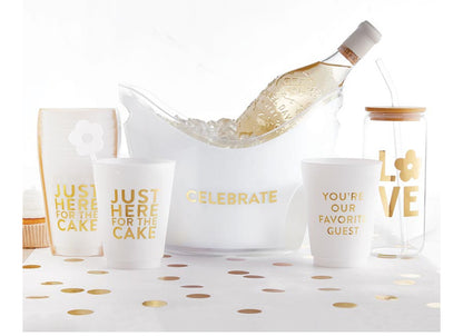 A bottle of white wine held by a clear ice bucket adorned with "CELEBRATE" in gold. Frosted Cups: “JUST HERE FOR THE CAKE” and “YOU'RE OUR FAVORITE GUEST.” Clear LOVE-themed tumbler. Sizes: N/A for wine bottle, cups, and tumbler.
