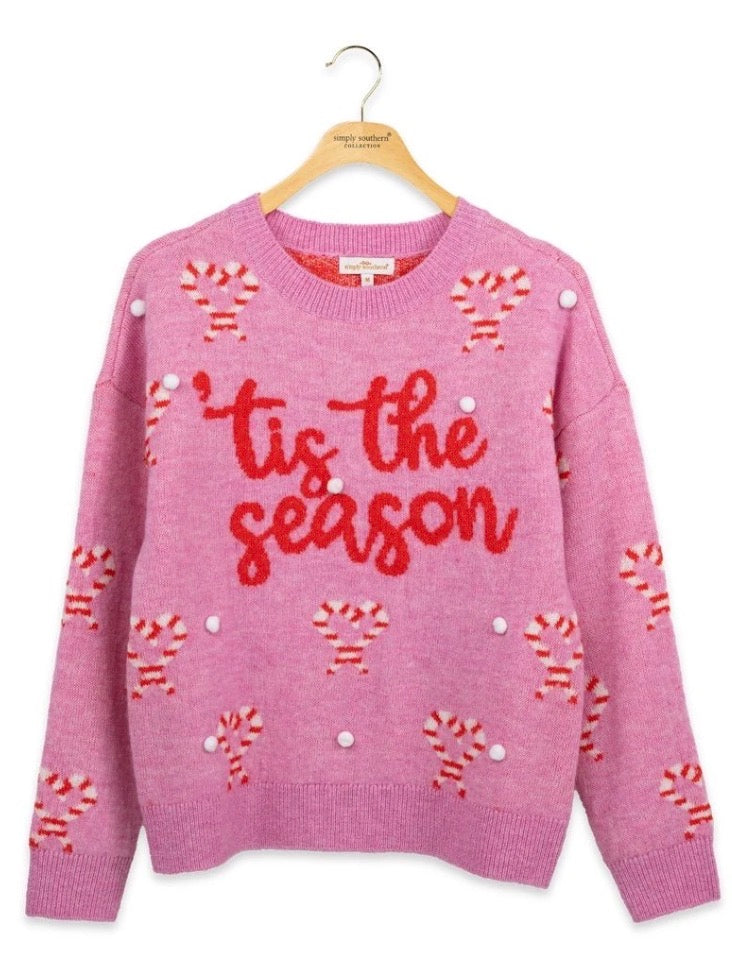 Red “‘tis the season” text, candy cane graphics, and white pompoms on a festive sweater. Available in sizes XS-XL.