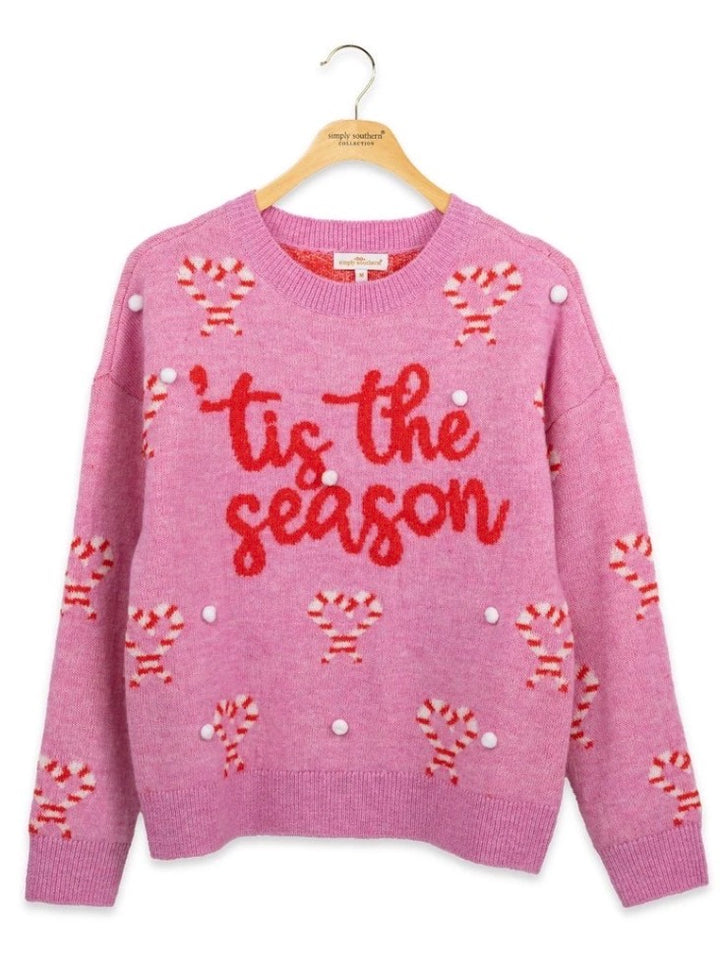 Red “‘tis the season” text, candy cane graphics, and white pompoms on a festive sweater. Available in sizes XS-XL.