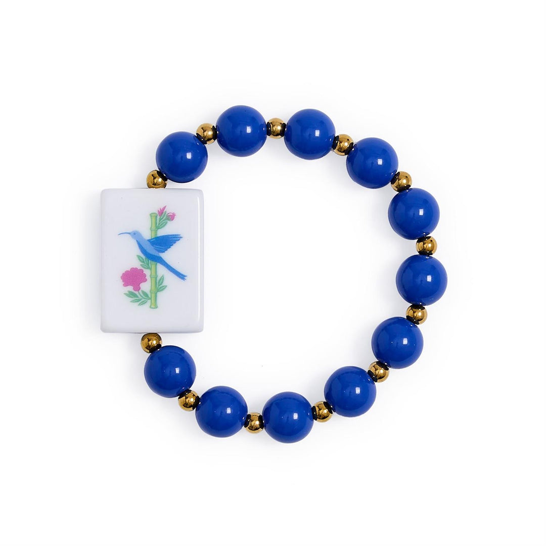 Mahjong Beaded Stretch Bracelet features alternating large blue and small gold beads, a rectangular white charm with a blue bird on a pink-flowered branch, and colorful acrylic Mahjong-inspired accents. Available in multiple sizes.