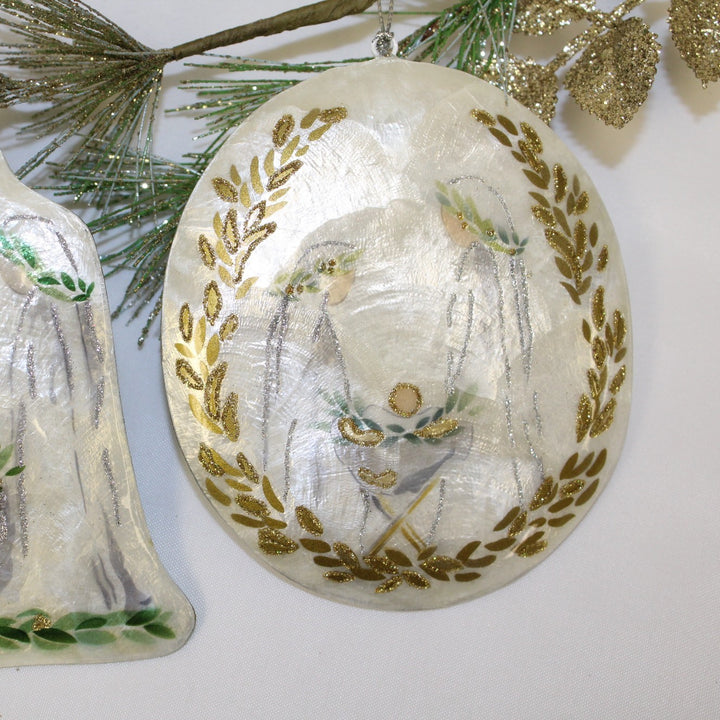 Handmade Holy Family Capiz Ornament: an oval iridescent shell with a painted Holy Family scene framed by a gold glitter laurel wreath.