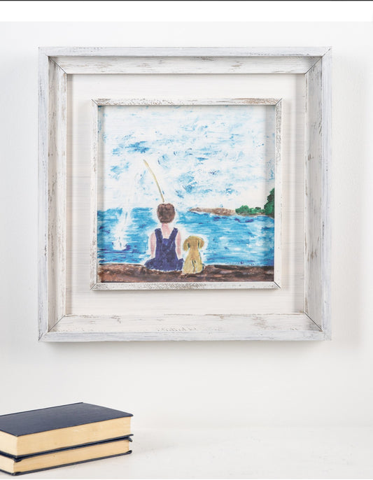 A Gone Fishing Wall Picture of a person and a dog sitting by a lake, displayed on a white wall above two stacked books. The frame is rustic white and complements the serene artwork. The background includes subtle