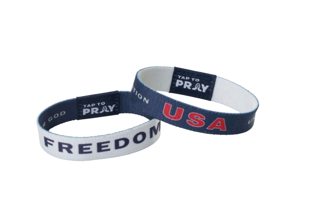 Two Tap To Pray™ Freedom wristbands: one white with blue “FREEDOM” text, one blue with red “USA” text, each with a logo tab.