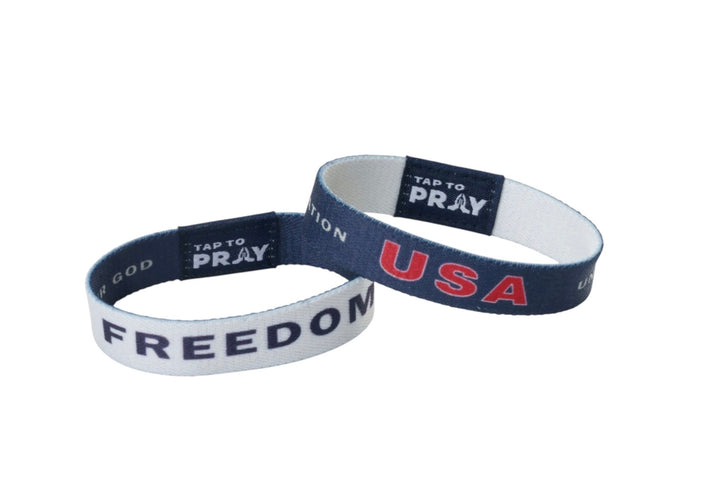 Two Tap To Pray™ Freedom wristbands: one white with blue “FREEDOM” text, one blue with red “USA” text, each with a logo tab.
